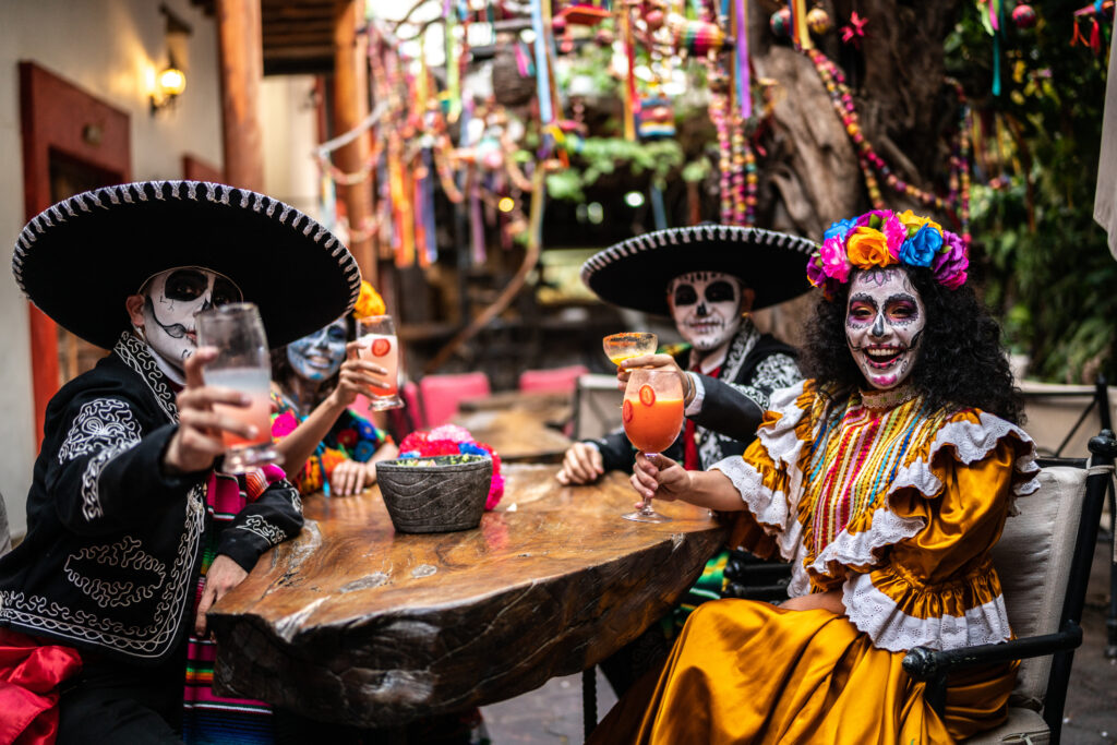 Group of 4 friends looking to camera doing a celebratory toast on the day of the dead at a bar, dressed in traditional Mexican dress for the festival, with faces painted